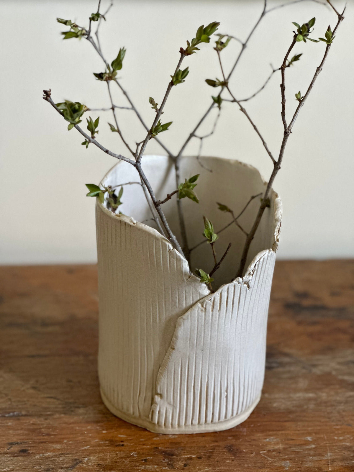 A handmade ceramic vase with a vertical linear impression and a rustic torn raw edge, displayed with some branches inside it.
