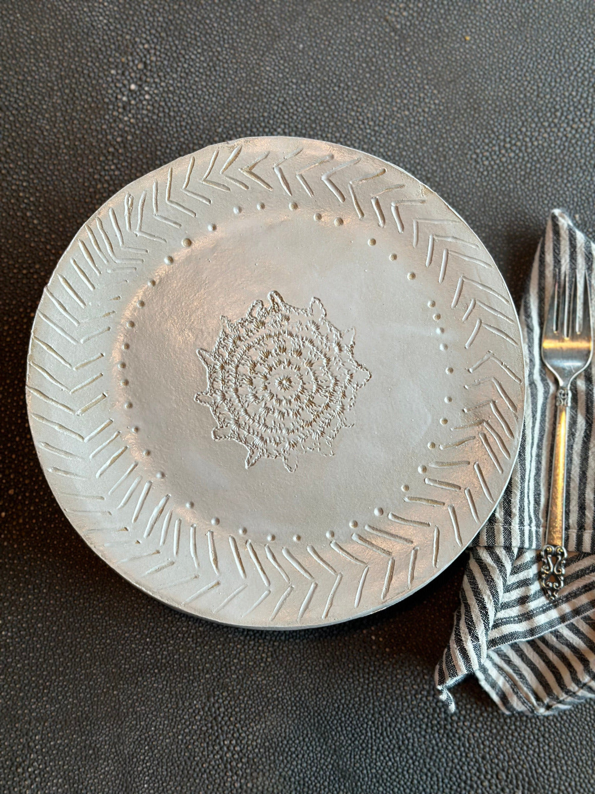 A matte white finish ceramic dinner plate with a geometric pattern around the rim and a floral design in the center, displayed on a dark surface alongside a striped napkin and a fork.
