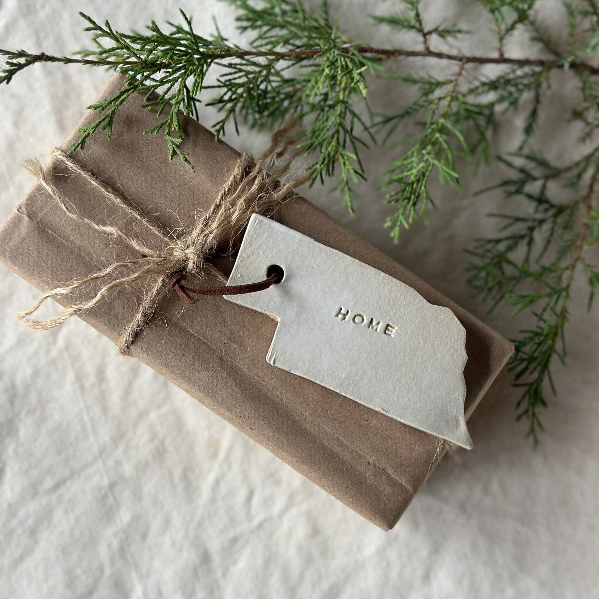 Handmade Nebraska 'home' Christmas ornament with a leather string, placed on a white surface surrounded by green pine needles.