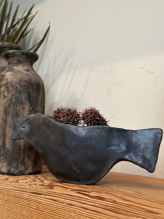 Black ceramic bird sculpture on a wooden surface with a textured vase and decorative items in the background.