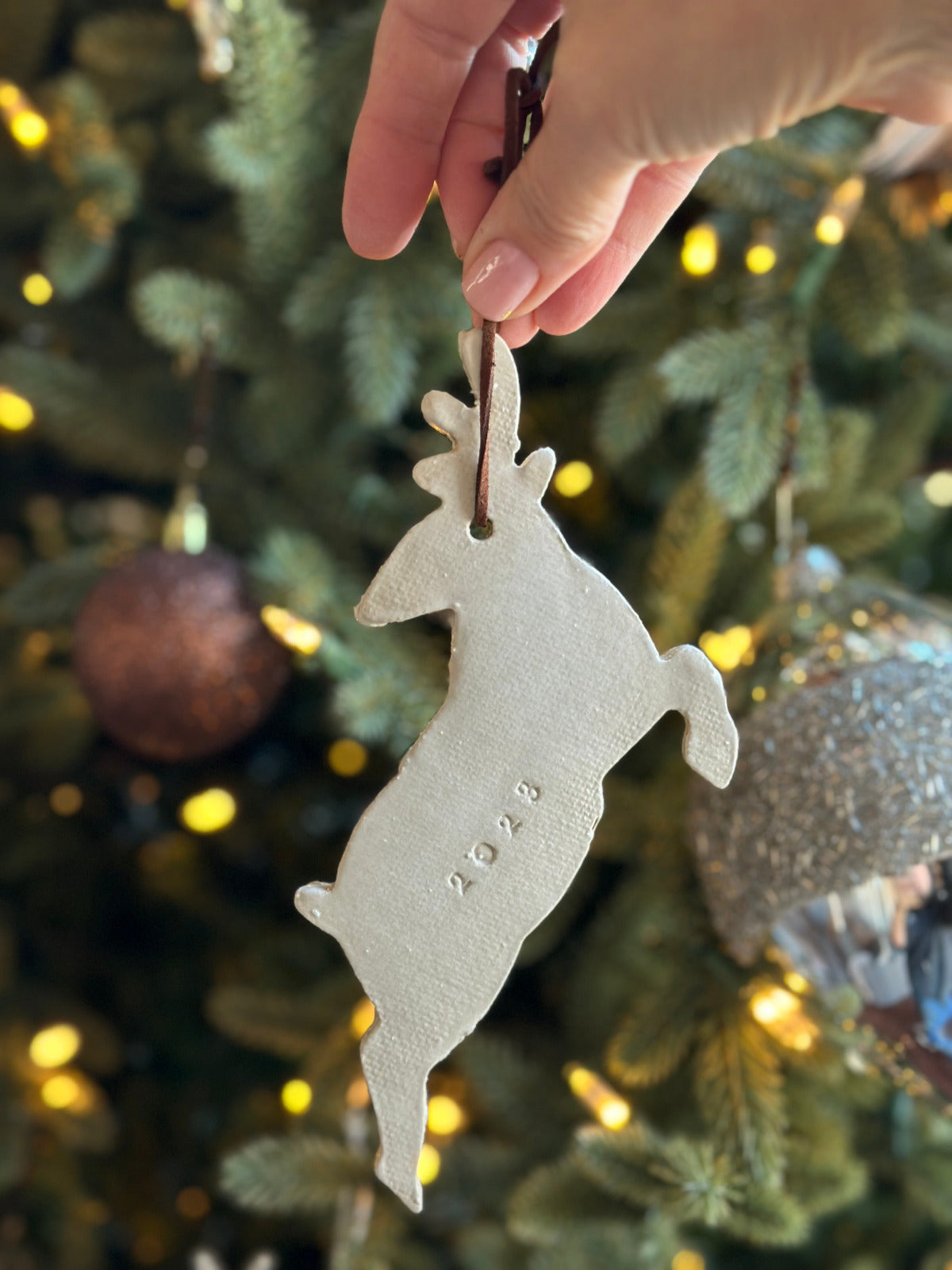 A hand holding a white reindeer-shaped Christmas ornament with a leather string, against a blurred Christmas tree background with lights.