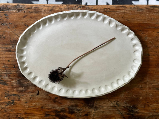 An oval ceramic plate with a matte white finish and thumbprint impressions along the edge, displayed on a wooden surface.