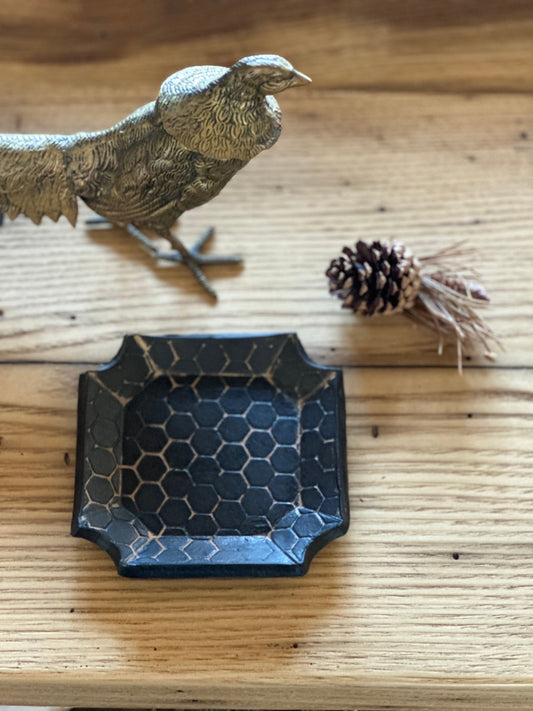 A matte black handmade trinket dish with a honeycomb impression, displayed on a wooden surface next to a bird figurine and some pine cones.