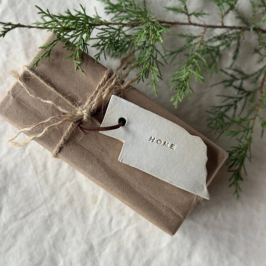 Handmade Nebraska 'home' Christmas ornament with a leather string, placed on a white surface surrounded by green pine needles.