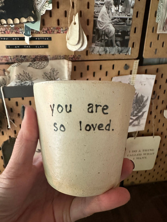 Hand holding a ceramic cup with 'you are so loved' text in front of a store display.