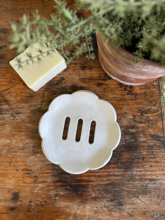 A matte white, scalloped edge ceramic soap dish with self-draining holes, displayed on a wooden surface.