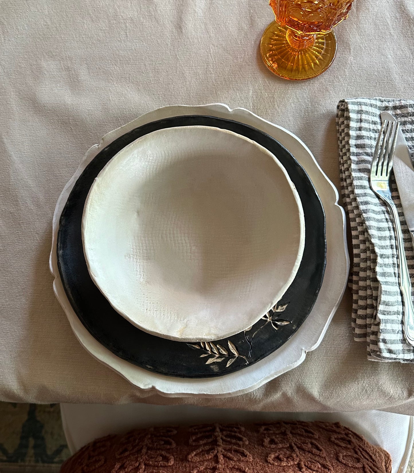 Set of handmade dishes including white charger, black salad plate with botanical impression and white matte bowl.