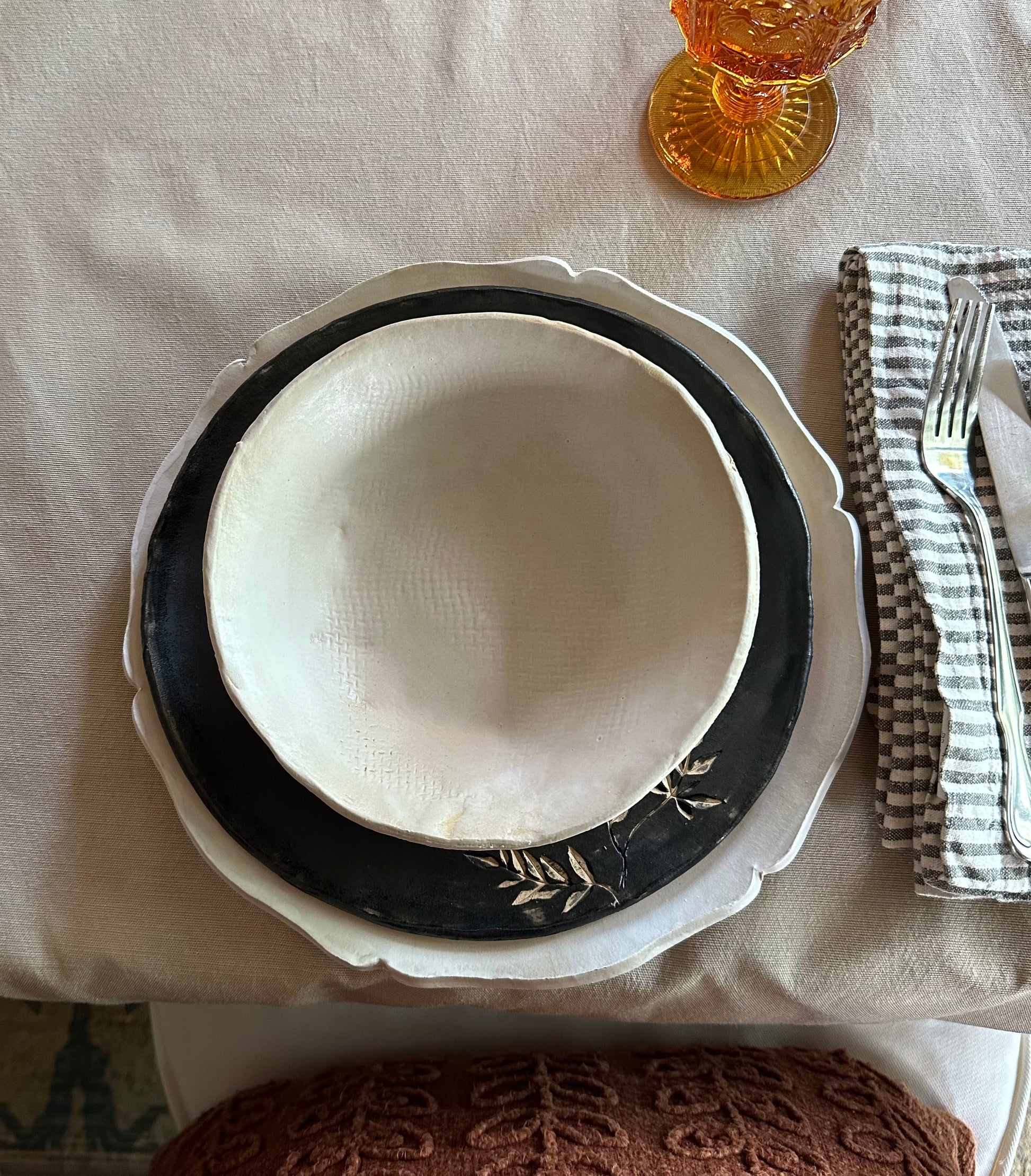 Set of handmade dishes including white charger, black salad plate with botanical impression and white matte bowl.