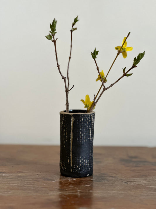 A ceramic bud vase with a black matte finish and burlap-like texture, containing branches with yellow flowers.