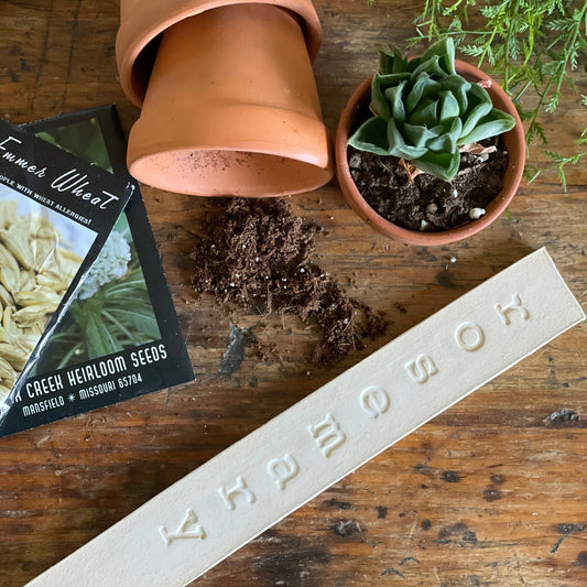 A brown ceramic garden marker with the text 'Farm Steady' on it, placed on a wooden surface next to a potted plant and some soil.