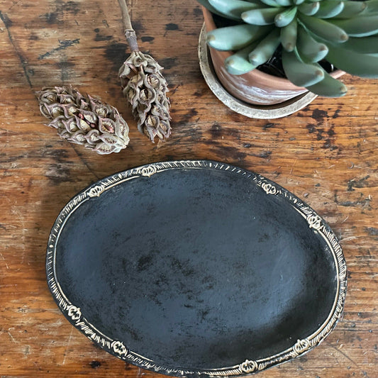 A black matte finish oval platter with decorative elements on the side, placed on a wooden surface with a potted plant in the background.