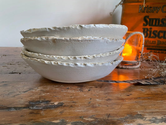 A set of three matte white stoneware bowls with a rustic edge finish on a wooden surface, with blurred lights and objects in the background.