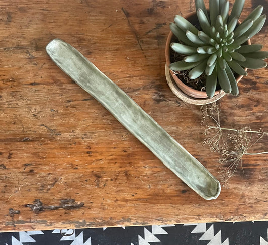 A handmade olive boat made of a green material, placed on a wooden surface, with a potted plant and decorative items in the background.